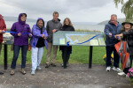 Eight people in wind-resistant clothing holding a blue ribbon in front of Stewart Park
